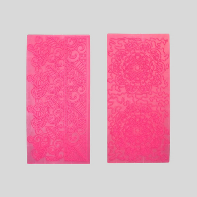 Two rectangular impression mats for fondant icing in a vibrant hot pink color, displayed side-by-side against a neutral light gray background. The left die features an intricate floral and scroll pattern, while the right die showcases a mandala design.