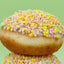 A glazed donut topped with pastel multicolor sprinkles, stacked on top of another donut against a solid light green background.