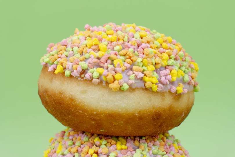 A glazed donut topped with pastel multicolor sprinkles, stacked on top of another donut against a solid light green background.