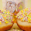 Two frosted donuts topped with pastel multicolor sprinkles, with a branded bag blurred in the background on a pale yellow surface.