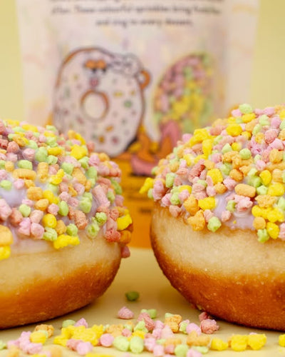 Two frosted donuts topped with pastel multicolor sprinkles, with a branded bag blurred in the background on a pale yellow surface.
