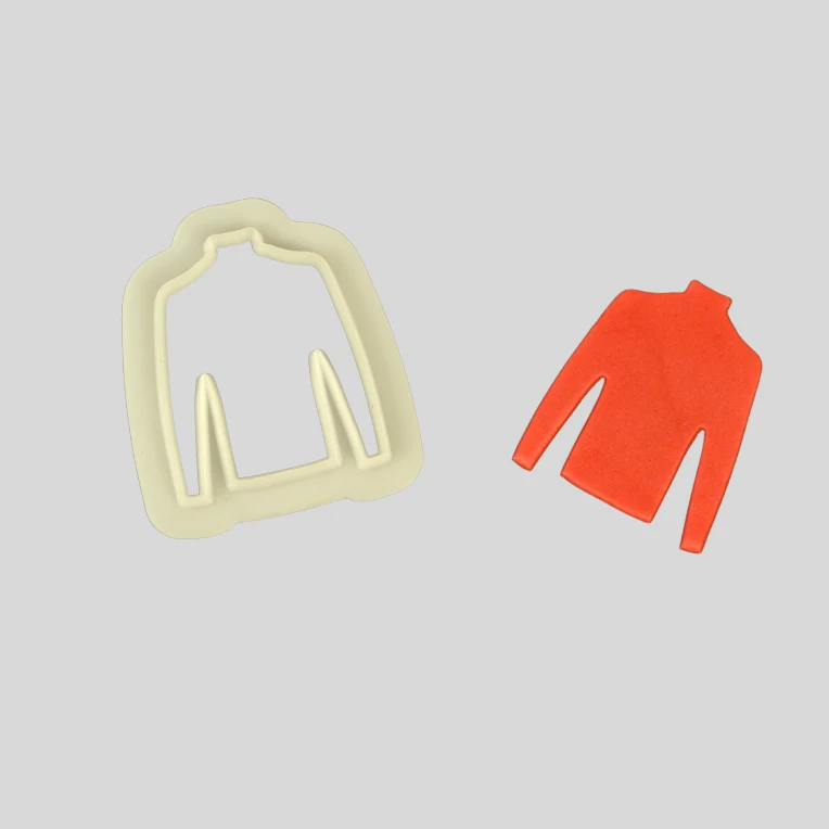 A cream-colored plastic jumper cutter shaped like a turtleneck sweater, shown next to a small orange-red molded piece of fondant on a solid gray background.