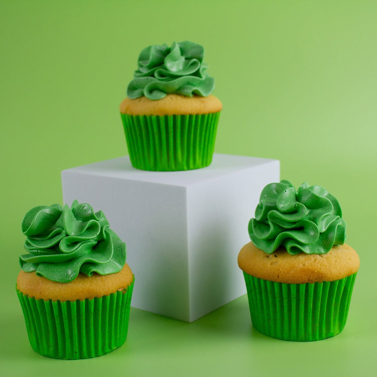 Three green-frosted cupcakes in bright green paper liners arranged around a white cube on a light green background.