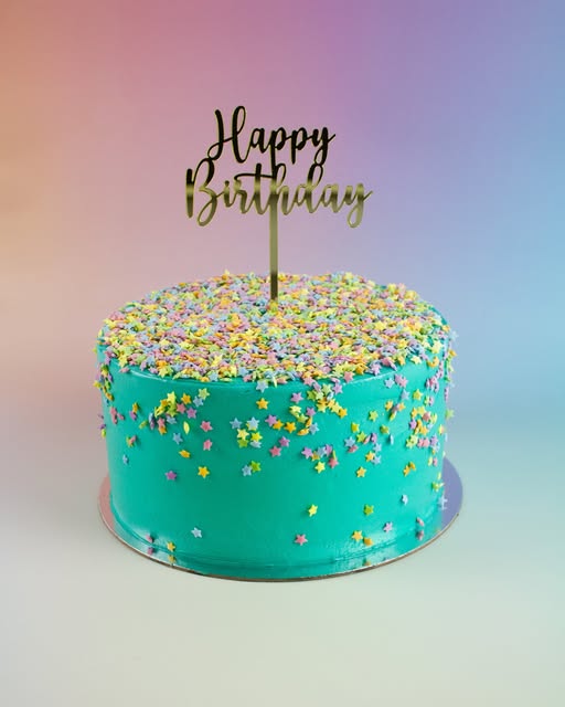 A round birthday cake with bright turquoise frosting, decorated with multicolored star-shaped sprinkles and topped with a gold 'Happy Birthday' script topper, set against a soft pastel gradient background.