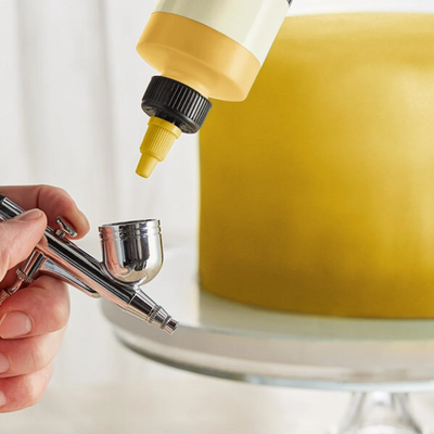 A close-up of a hand holding a silver airbrush tool while a yellow squeeze bottle dispenses yellow material onto a round yellow cake on a white cake stand.
