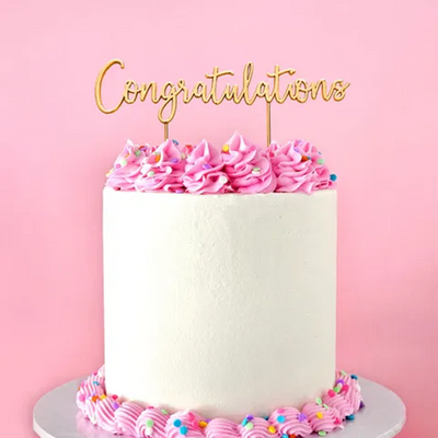 White cake with pink frosting and 'Congratulations' topper on a pink background