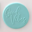 A round light blue fondant plaque featuring the words 'Good Vibes' in a cursive script, set against a plain light pink background.