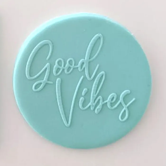 A round light blue fondant plaque featuring the words 'Good Vibes' in a cursive script, set against a plain light pink background.