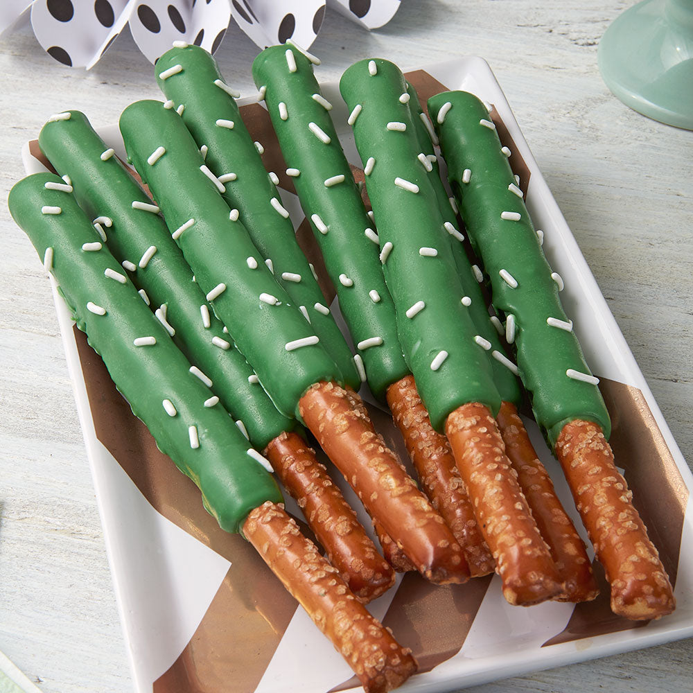 A set of green chocolate-covered pretzel sticks with white sprinkles, arranged on a white rectangular serving tray with brown paper liner.