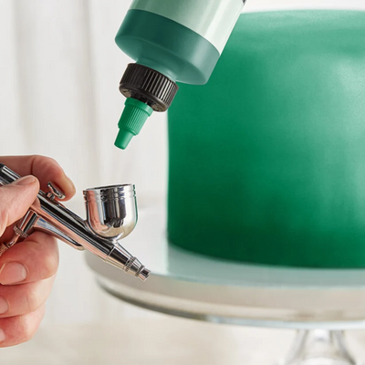 A close-up of a hand holding an airbrush with a green color bottle pointed at a green cake on a stand, set against a soft white background.