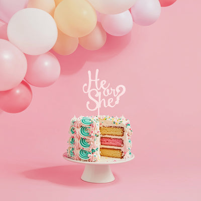 Decorative cake with 'He or She?' topper on a pink background with balloons