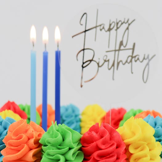 A close-up of a colorful birthday cake with piped frosting rosettes in orange, green, red, yellow, and blue, three lit blue candles, and a clear 'Happy Birthday' script topper on a light gray background.
