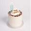 A round birthday cake with white frosting, chocolate crumb decoration around the top edge, three light blue candles, and a white 'Happy Birthday' topper on a silver cake board against a light gray background.