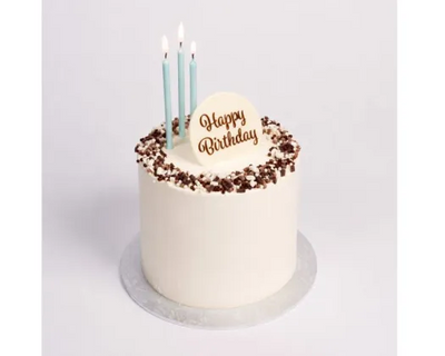 A round birthday cake with white frosting, chocolate crumb decoration around the top edge, three light blue candles, and a white 'Happy Birthday' topper on a silver cake board against a light gray background.
