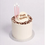 A round birthday cake with white frosting, chocolate sprinkles around the rim, three pink candles, and a white 'Happy Birthday' plaque on top, set on a silver cake board against a light gray background.