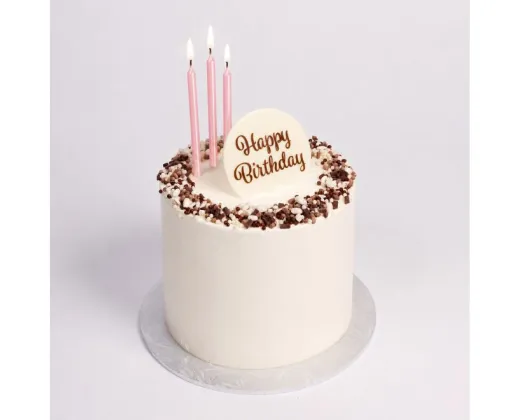 A round birthday cake with white frosting, chocolate sprinkles around the rim, three pink candles, and a white 'Happy Birthday' plaque on top, set on a silver cake board against a light gray background.