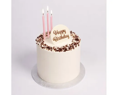A round birthday cake with white frosting, chocolate sprinkles around the rim, three pink candles, and a white 'Happy Birthday' plaque on top, set on a silver cake board against a light gray background.
