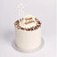 A single-tier white frosted birthday cake with chocolate sprinkles around the top edge, three white candles, and a round white topper that reads 'Happy Birthday' in brown lettering, shown on a light gray background.