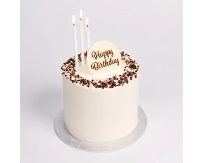 A single-tier white frosted birthday cake with chocolate sprinkles around the top edge, three white candles, and a round white topper that reads 'Happy Birthday' in brown lettering, shown on a light gray background.