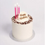 A white frosted birthday cake with chocolate sprinkles around the edge, three pink candles, and a white topper that reads 'Happy Birthday' in gold lettering, shown on a light gray background.