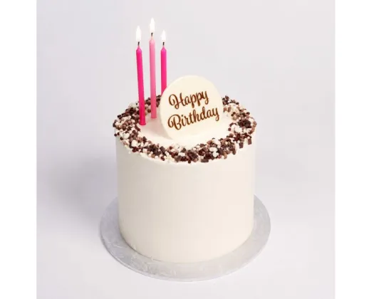 A white frosted birthday cake with chocolate sprinkles around the edge, three pink candles, and a white topper that reads 'Happy Birthday' in gold lettering, shown on a light gray background.