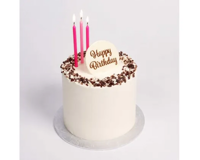 A white frosted birthday cake with chocolate sprinkles around the edge, three pink candles, and a white topper that reads 'Happy Birthday' in gold lettering, shown on a light gray background.