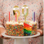 A person holding a frosted rainbow birthday cake with sprinkles and large gold number 60 candles on a white cake stand.