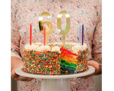A person holding a frosted rainbow birthday cake with sprinkles and large gold number 60 candles on a white cake stand.