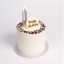 A single round birthday cake with white frosting, chocolate crumb decoration around the top edge, a white heart-shaped topper with 'Happy Birthday' in gold script, and three lit silver candles, shown on a light gray background.