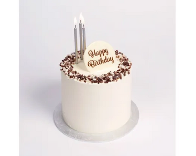 A single round birthday cake with white frosting, chocolate crumb decoration around the top edge, a white heart-shaped topper with 'Happy Birthday' in gold script, and three lit silver candles, shown on a light gray background.