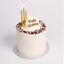 A round birthday cake with white frosting, chocolate crumb topping, and three gold taper candles on a white background. The top features a white plaque with 'Happy Birthday' written in gold script.