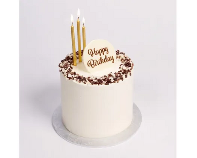 A round birthday cake with white frosting, chocolate crumb topping, and three gold taper candles on a white background. The top features a white plaque with 'Happy Birthday' written in gold script.