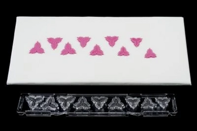 A white rectangular tray or mat with a row of small pink fondant icing holly leaves across the top, shown above a clear plastic cutter for the same shapes