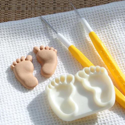 A baby footprint cutter mould by Jem set on a white textured mat, featuring two small beige baby footprints, alongside two yellow-and-white carving tools.