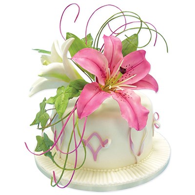 A decorated white cake with pink and white lilies, green leaves, and purple curly vine accents on a white background.
