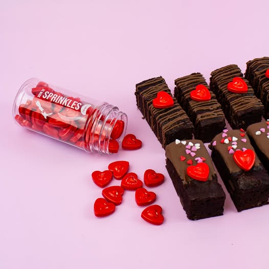 A clear plastic jar labeled 'The Sprinkles' filled with red heart-shaped candies, shown tipped over with several red heart sprinkles spilled out beside rows of chocolate brownies decorated with red and pink heart candies on a light pink background.