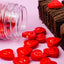 Red heart-shaped candy pieces spilled from a clear plastic jar next to chocolate brownies decorated with red heart candies, on a pink background.