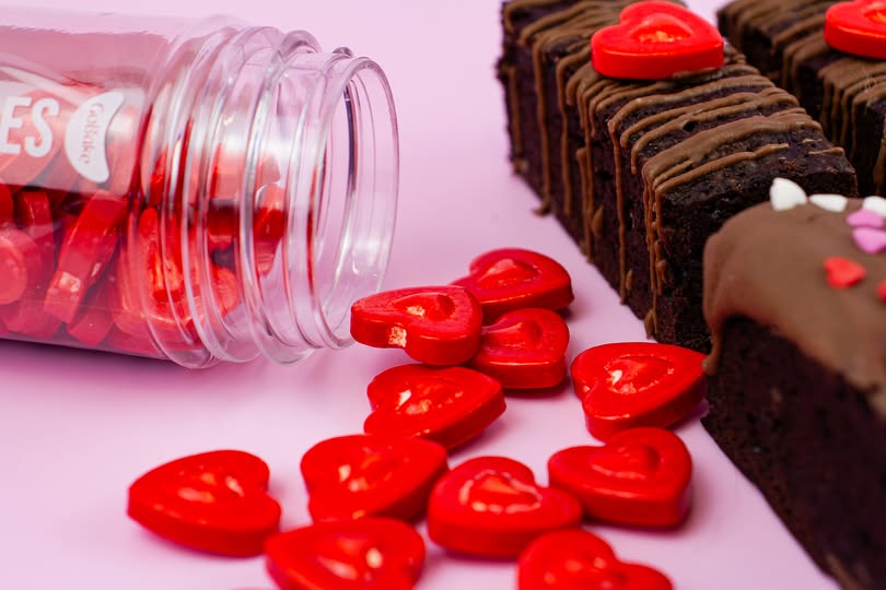 Red heart-shaped candy pieces spilled from a clear plastic jar next to chocolate brownies decorated with red heart candies, on a pink background.