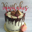 A frosted cake with chocolate drip, pink rosebuds, two brown macarons on top, and a pink macaron beside it. A gold and pink 'KiwiCakes' script topper stands on the cake, and the cake sits on a gold board against a white brick wall background.