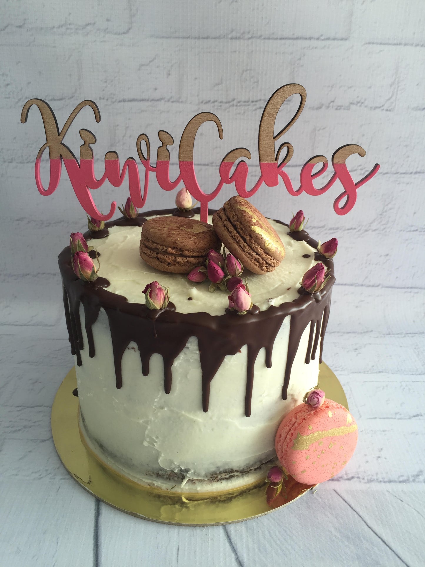 A frosted cake with chocolate drip, pink rosebuds, two brown macarons on top, and a pink macaron beside it. A gold and pink 'KiwiCakes' script topper stands on the cake, and the cake sits on a gold board against a white brick wall background.