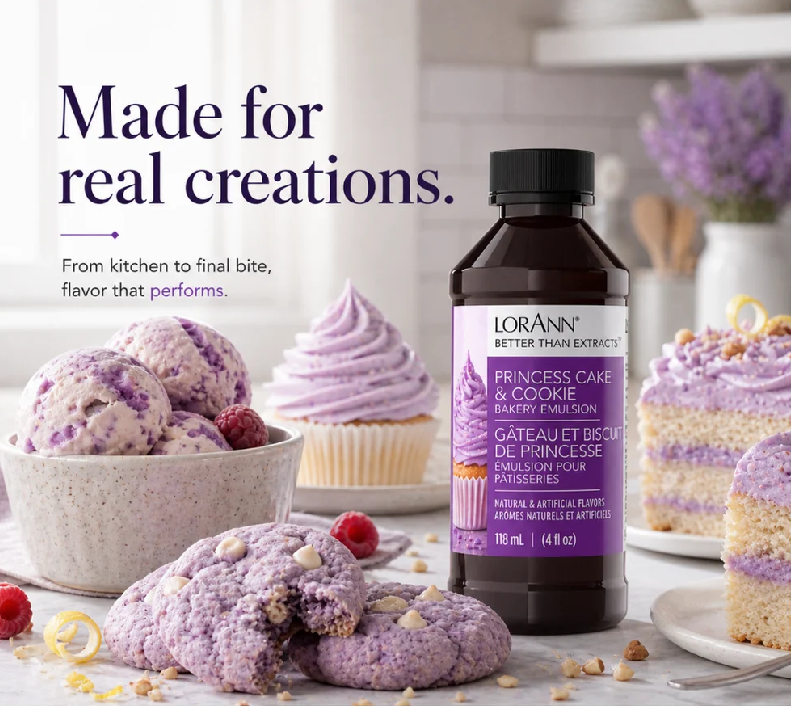 A bottle of Lorann's Better Than Exctract Cake & Cookie Emulsion stands on a marble surface next to purple berry-topped cookies, a cupcake, and ice cream scoops in a bright kitchen setting. Large text on the left reads 'Made for real creations.