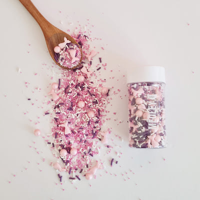 Pink and purple sprinkles on a wooden spoon with a container of sprinkles on a light background