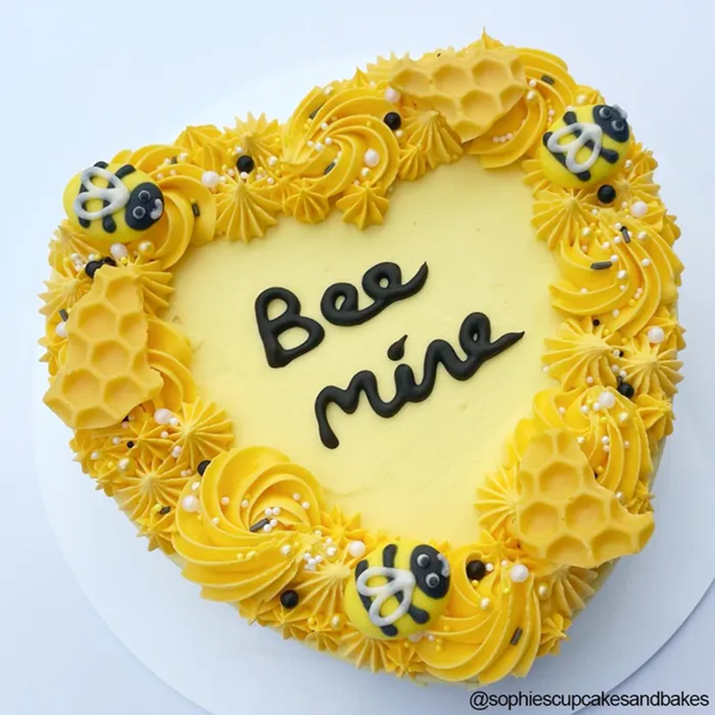 Heart-shaped cake with yellow frosting, bees, and 'Bee mine' text on a white background