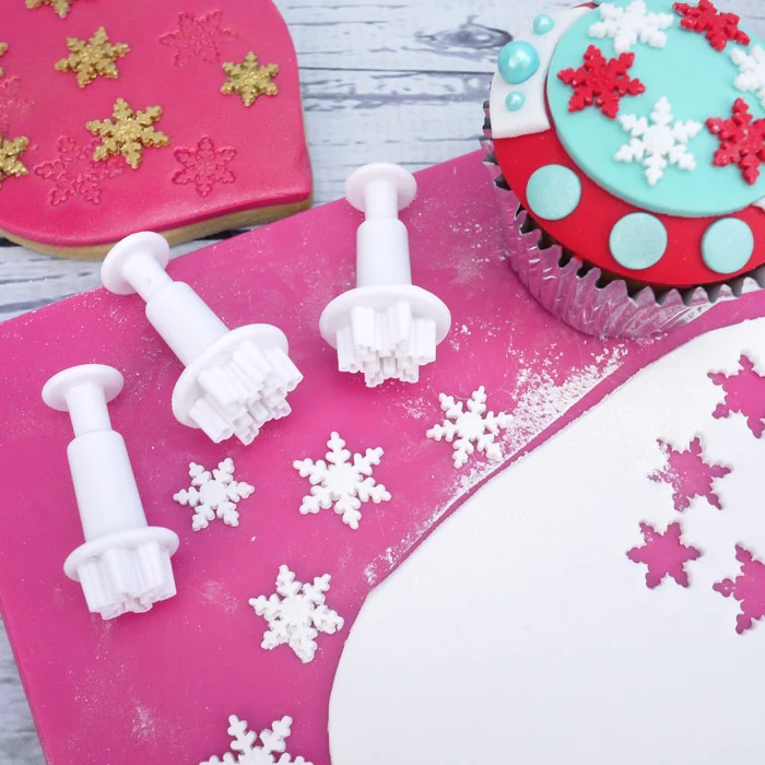 Snowflake cookie cutters pink fondant icing surface with snowflakes and a decorated cupcake.