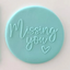 A round light blue fondant plauqe with the words 'Missing you' and a small heart in matching light blue icing, shown on a plain light background.