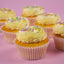 A close-up of several frosted cupcakes arranged on a pink background. The cupcakes have pale yellow whipped frosting topped with multicolored round sprinkles and are in white paper liners.
