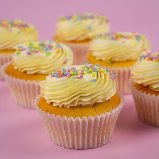 A close-up of several frosted cupcakes arranged on a pink background. The cupcakes have pale yellow whipped frosting topped with multicolored round sprinkles and are in white paper liners.