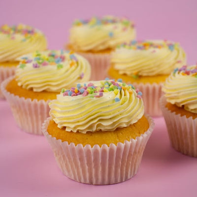 A close-up of several frosted cupcakes arranged on a pink background. The cupcakes have pale yellow whipped frosting topped with multicolored round sprinkles and are in white paper liners.