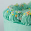 A close-up of a mint green frosted cake with piped rosette borders decorated with colorful round sprinkles, set against a light gray background.