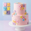 Two-tier pink cake decorated with colorful flowers on a white stand against a light purple background.